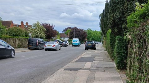 A street in Northampton, with several cars on it, parked and being driven. A path is to the right, with patches on it, with bushes and trees down the right-hand side. There are houses to the left, with more trees and bushes and more parked cars.