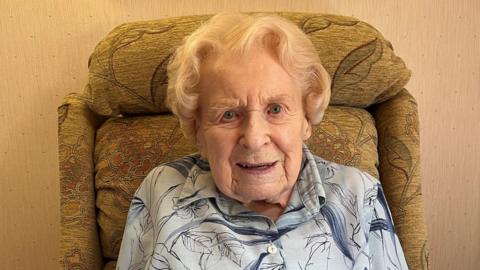 Edith Roberts. A woman sits in a chair. She has short blonde hair and is smiling