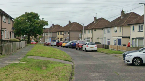 Whinsmoor Drive in Carlisle. Beige semi-detached houses line either side of a curving road which has grass verges and parked cars down one side.