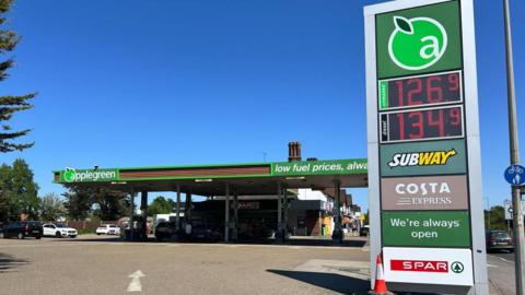 Image of a petrol station forecourt. There are red cars next to the pumps. Neighbouring houses can be seen. The petrol station is covered by a canopy bearing the words "applegreen" and "low fuel prices, always". 