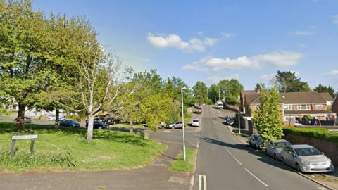 The turning for Walton Drive, with a street name sign on the left that is on top of a grassy area with trees. On the right are parked cars on a verge, a tree and some housing. The road goes up a slight incline into the distance.