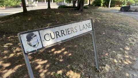 A road sign at the edge of a residential street. The sign reads Curriers Lane. There are trees behind the sign and a grass area. A lone car is parked further up toward the right. 
