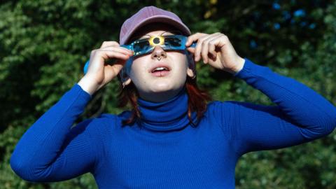 A woman wearing blue looking through special cardboard glasses for looking at the sun