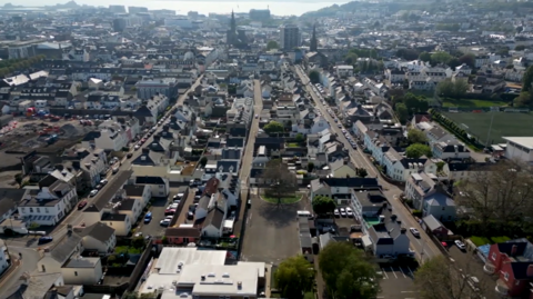 Aerial view of a densely populated town with rows of houses and buildings stretching towards the coastline. The streets are lined with parked cars, and a few green spaces are visible within the urban landscape.