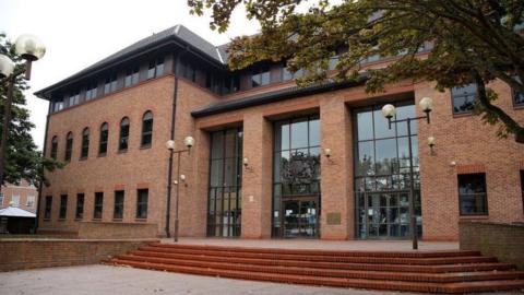 An exterior view of Derby Crown Court, a red brick three-storey building with brown window and door frames and steps leading up to it.