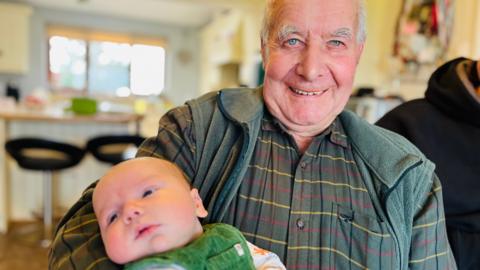 Bob holding his great-grandson, who is eight weeks old. Bob has a green shirt with thin yellow, red and blue horizontal lines and a green gilet. He has short white hair and is bald on top. The baby has a green outfit on