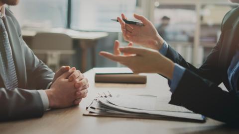 Close up image of two people sitting at a desk, facing each other and gesturing with their hands, one person holds a pen and has a clipboard