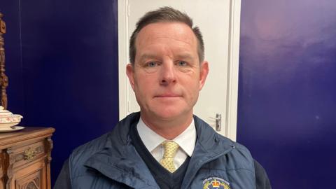 A man with short brown hair wears a navy v-neck sweater, navy gillet, white shirt and yellow tie. Behind him is a white door which splits a purple wall.