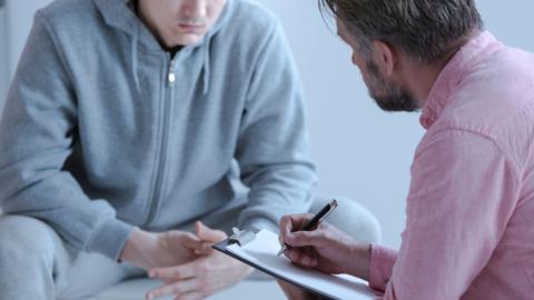 A stock photo of a psychologist who is wearing a pink shirt interviewing his patient who is in a grey tracksuit during a therapy Counselling Session