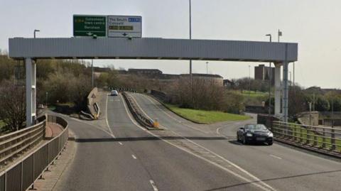 A view of the bridge just before the crash site. There are four lanes, two in each direction, but no barrier between them until halfway up the slope. An overhead gantry signals which lanes go where.