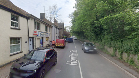 A Google Street view image of a two-laned road with a house and comunity store on one side and a wall and trees on the other side. It is a two-lane road which is made quite narrow by parked cars.