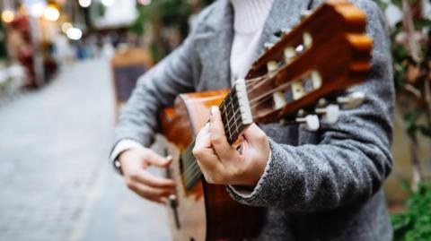 Generic image of a woman playing acoustic guitar by the side of a pedestrianised street with a blurred background of street cafe seating and buildings with lights showing. She is wearing a grey jacket and white polo-neck. Her face is not visible.