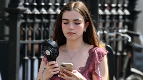 A young girl in a pink top with brown hair walks along a street carrying a portable black fan, whilst looking down at her phone in a clear case. 