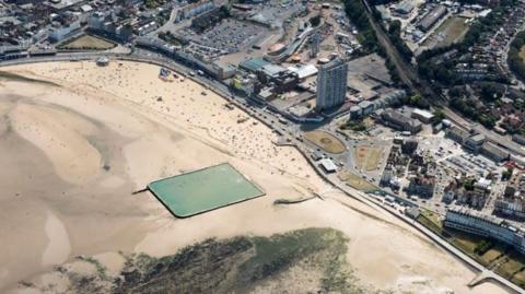 A rectangular pool of green seawater on a beach, surrounded by sand as the tide is out