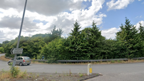 A roundabout with lots of large trees and a sign reading "Town centre, Leisure centre, Chawson" on the left hand side. A Citroen car can be seen driving round.