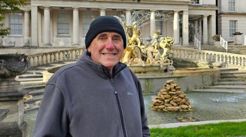 A man in his 70s smiles as he stands in front of an ornate water fountain at the front of a large Regency-style building. He is wearing a black beanie and a grey zip-up fleece. There are steps framing the fountain, which has statues looking over it and a pile of stones in the middle of the water.