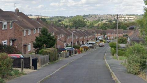 A mid-20th century residential street runs downhill with red-brick, semi-detached houses to either side and more at the the bottom of the hill. Cars are parked in driveways and some by the side of the road. In the background hills rise from a valley with more homes visible in the distance. 