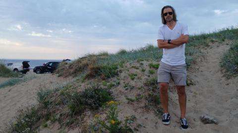 Councillor Dr Tristan Learoyd standing on sand at Marske beach. He has long hair and is wearing sunglasses, a white T-shirt and beige shorts with blue trainers.