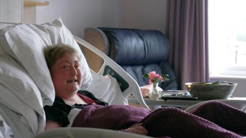 A woman with short brown hair lies in a clinical bed in a raised position. She is surrounded by pillows and covered in a burgundy blanket. In the background a table with fruit bowl and flowers and a navy armchair next to a window are visible.