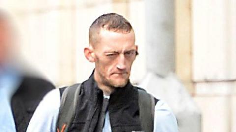 Craig Rowland - a still of a man with short brown hair wearing a light blue collared shirt, a skinny grey tie and a black vest jacket. He has one eye closed and is looking at the camera as he walks near a white stone building. A person's face in the background is blurred.