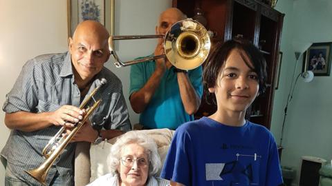  Marcus Fernando, left, wearing a blue and white striped shirt and holding a trumpet. Paul Fernando, centre, wearing a blue shirt and holding a trombone. 
 Phyllis Fernando, in the middle of Marcus and Paul, sat on a cream chair, with short white hair and glasses. Sebastian Fernando, front right, wearing a blue shirt and looking directly at the camera. 