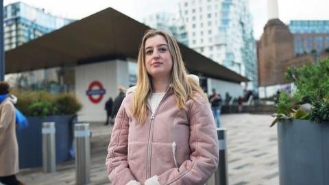Beth, in a light pink coat, stands at the entrance of the Battersea Power Station Underground station in London. Behind her are modern glass buildings and the tall white chimneys of the redeveloped Battersea Power Station.