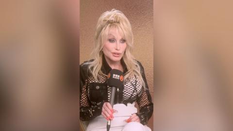 Dolly Parton wearing black mesh shirt speaks into a BBC microphone during sitdown interview