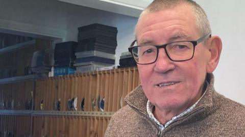 Steve Webber poses behind a desk holding a DVD. There are more DVDs piled on the counter. He is wearing glasses, has cropped grey hair and has on a brown pullover.