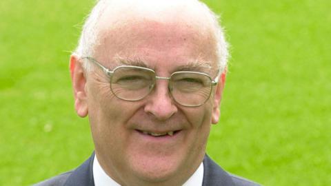 Former Colchester United and EFL chairman Peter Heard standing on the grass of a football pitch smiling while wearing glasses and a dark jacket and white shirt