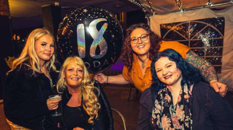 Bayley and three other women at a birthday party in a marquee. One of the women is holding a balloon marked "18 today"