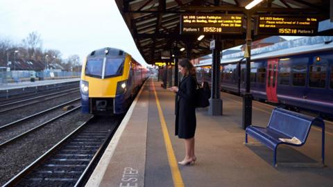 A woman is on her phone while she waits on a train platform. There is a yellow and blue train on the tracks in front of her