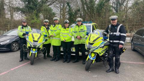 Several officers from the Tasking Team posing for a picture. A police car and two police motorcycles are beside them. It is an overcast day.