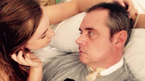 Simon Dobbin lying in a hospital bed, looking his daughter in the eye. She is holding his head and leaning towards him.