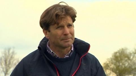 A TV still image of Duncan Farrington in a navy jacket with a red trim on the centre zipper. He has brown hair and is standing in an outdoor setting.