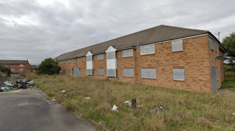 An exterior view of Admiral Court Nursing home. The building is a long brick structure with windows with white window frames. 