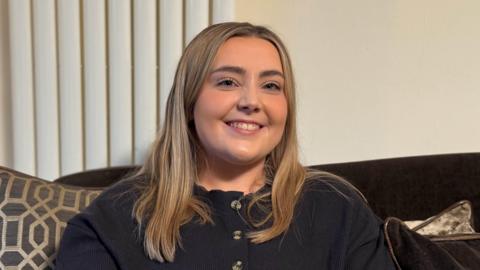 Kelsey is sitting on a brown velvet couch and is smiling down the lens. She has straight blonde hair which sits just below her shoulders. She is wearing a navy cardigan with tortoiseshell buttons.