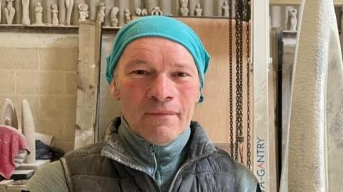 Nicolas Moreton is looking at the camera. He is wearing a turquoise head covering and a blue top and black gilet jacket. He is standing in a studio and there are stone sculptures behind him.