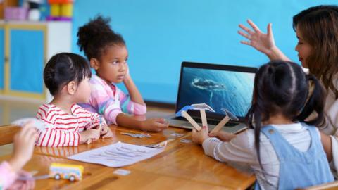 Three young children sitting round a desk in a school environment with a laptop on the table. A teacher has her hand raised and is expressing herself.