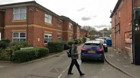 A Google Maps view of Lancaster Close in Woking. There is a person walking across a road, with parked cars on one side. 