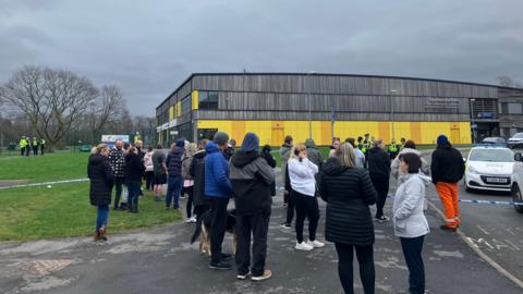 A group adults who look like they could be parents standing behind blue and while police tape across a foot path and road towards a two storey wood and yellow clad school building. About sixt police officers in high viz jackets can bee seen in a groups beside a police car about half way to the school building. Another group of four officers are together at the side entrance of the school building. It's a grey day. 