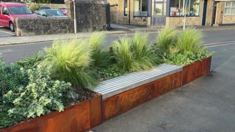 A planter with rusty walls has been completed, green plants sway in the wind and a bench for is on one side. 
