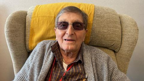 Nigel Williamson wearing dark sunglasses sat in a chair wearing a grey cardigan and dark checked shirt