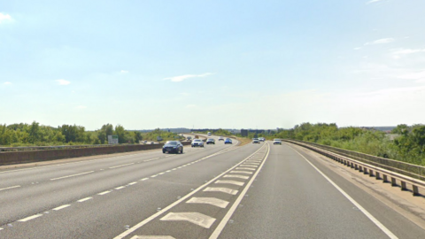 A general view of the A421 dual carriageway near Kempston in Bedfordshire. Numerous cars can be seen travelling on the carriageway while a car enters the road via a slip road. A bright blue sky can be seen above with trees and bushes lining the road.