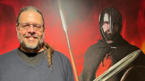 Brett Thorn standing in front of a life-size photograph of a man dressed as a Saxon warrior. Brett is on the left and has greying hair pulled back into a pony tail over his left shoulder. He is wearing glasses and holding up a replica sword, whose blade is pointing to the right.