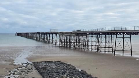 The iron pier runs from the right to the left down a sandy beach into the sea in the distance. There is a large cluster of rocks on the sand to the left. It is a cloudy but fairly bright day and the sea and very calm.