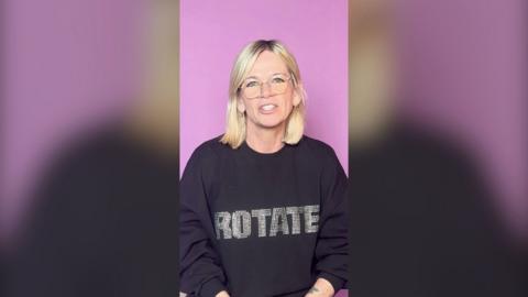 Zoe Ball, with blonde hair and glasses, in black jumper against a pink wall.