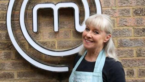 A smiling Penny Hester posing in front of the MasterChef logo mounted on a brick wall. Penny has long tied back white hair with a fringe and she is wearing a dark top with a light blue apron. The logo is a white letter M surrounded with a spiral.