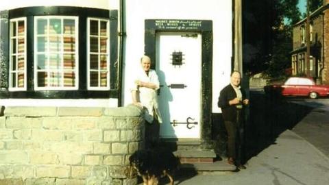An old colour photo of two men outside a corner shop. The building is painted white and has three steps leading up to a white painted door with a black door frame. To its left is a bow window with black window frames. Below the window is a stone wall. One man dressed in a white overall stands with a hand on the wall next to the door. In front of him him is a brown and black coloured dog. On the right is a man wearing dark trousers and cardigan. Both men appear to be of middle age and are looking towards the camera and smiling.
