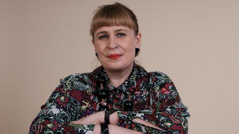 A woman with blonde hair tied back. She is wearing a multi-coloured patterned top and has her arms folded.