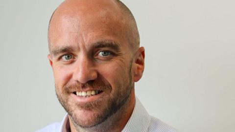 A bearded balding man with a blue checked shirt.